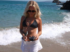 Woman in a black bikini and white skirt standing on a beach with ocean waves in the background